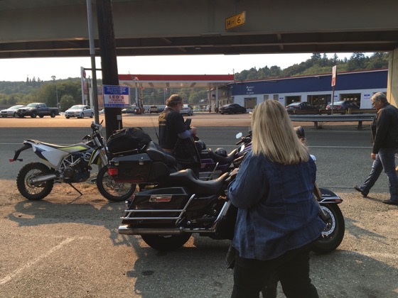 A 2016 Husqvarna 701 Enduro parked next to a Harley-Davidson bagger at a gas station with people standing around