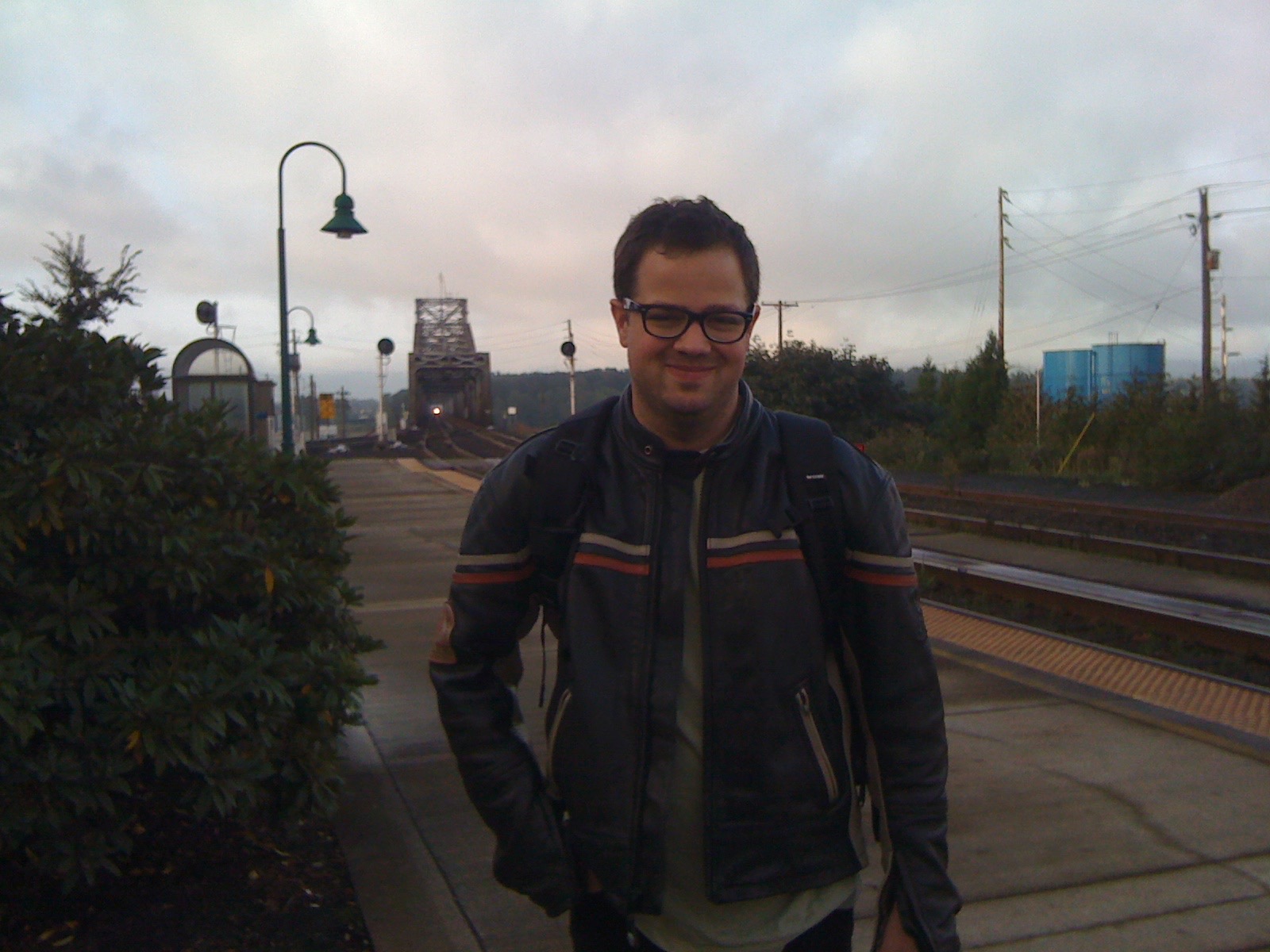 Jonathan in motorcycle gear on a train platform with a railroad bridge behind